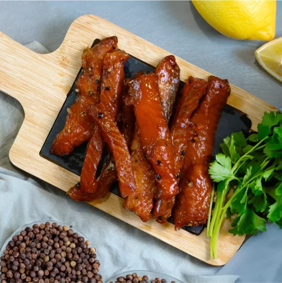Image of candied peppercorn salmon strips, gleaming with glaze, on a wooden board. Slices of fresh lemon and a small glass bowl of whole peppercorns are arranged alongside, hinting at the flavor profile of the dish.