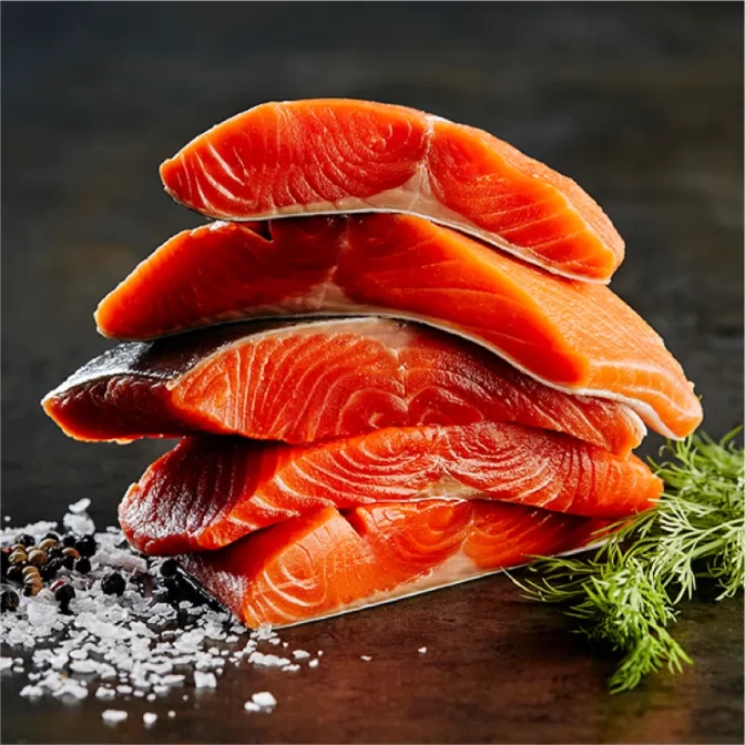 Image displays a stack of five raw wild Sockeye salmon fillets on a dark table. Accompanied by sprigs of fresh dill, chunks of coarse salt, and scattered peppercorns, suggesting the seasoning for the preparation.